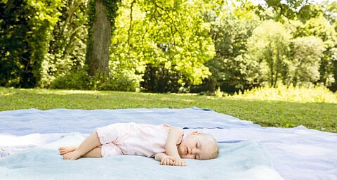 Egy kisbaba fekszik egy takarón egy füves területen, amelyet fák vesznek körül.