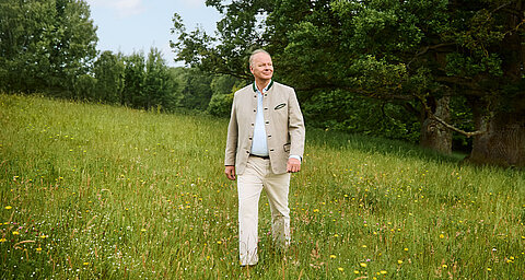 A man in a beige jacket and white pants walks through a grassy field with yellow flowers.