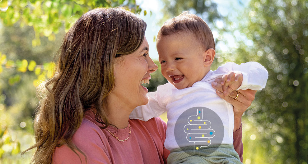 A woman in a pink shirt holds a laughing baby in a white shirt, both surrounded by greenery.