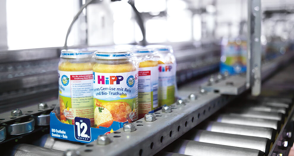 A conveyor belt in a factory setting transports transparent bottles with yellow contents, labeled with the word "HiPP" and other text.