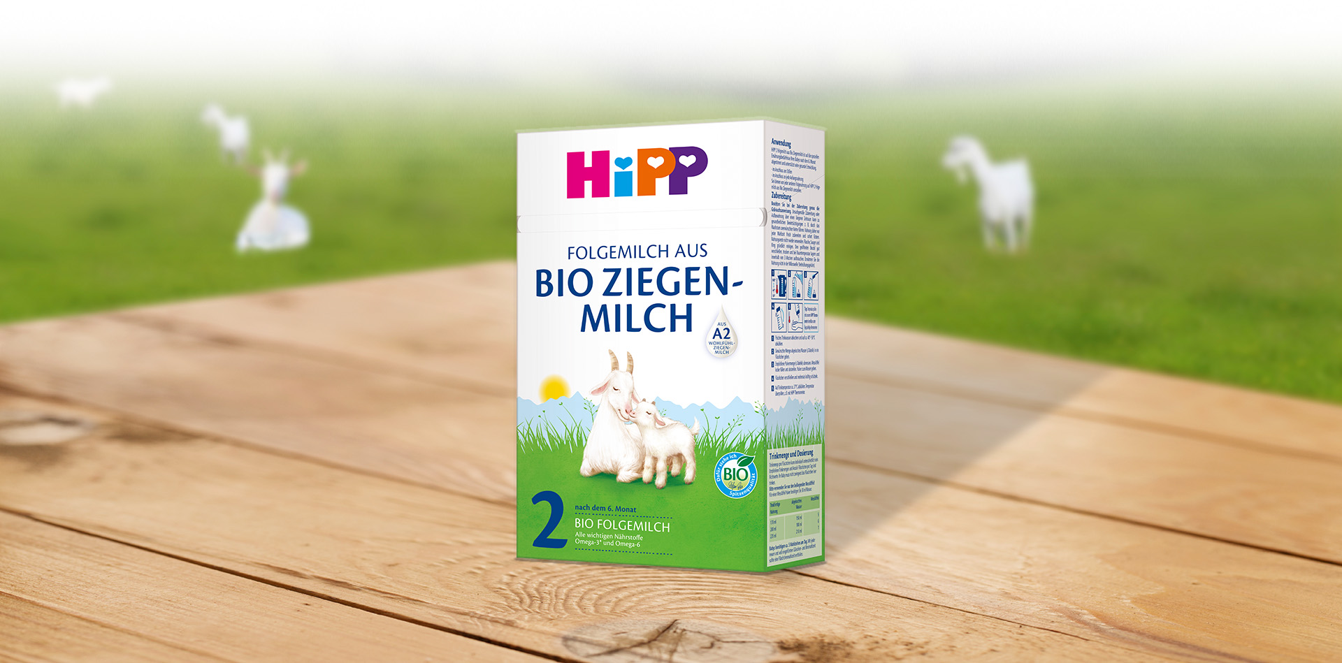 Egy fafelületre helyezett doboz Hipp bébiétel, 2. fokozat, kecske illusztrációval és zöld mezővel a háttérben.