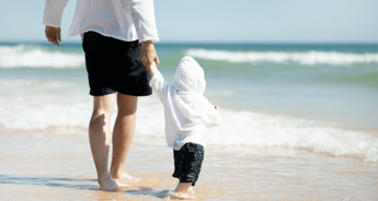 Egy személy és egy gyermek kéz a kézben sétál a strand szélén, a háttérben az óceán.