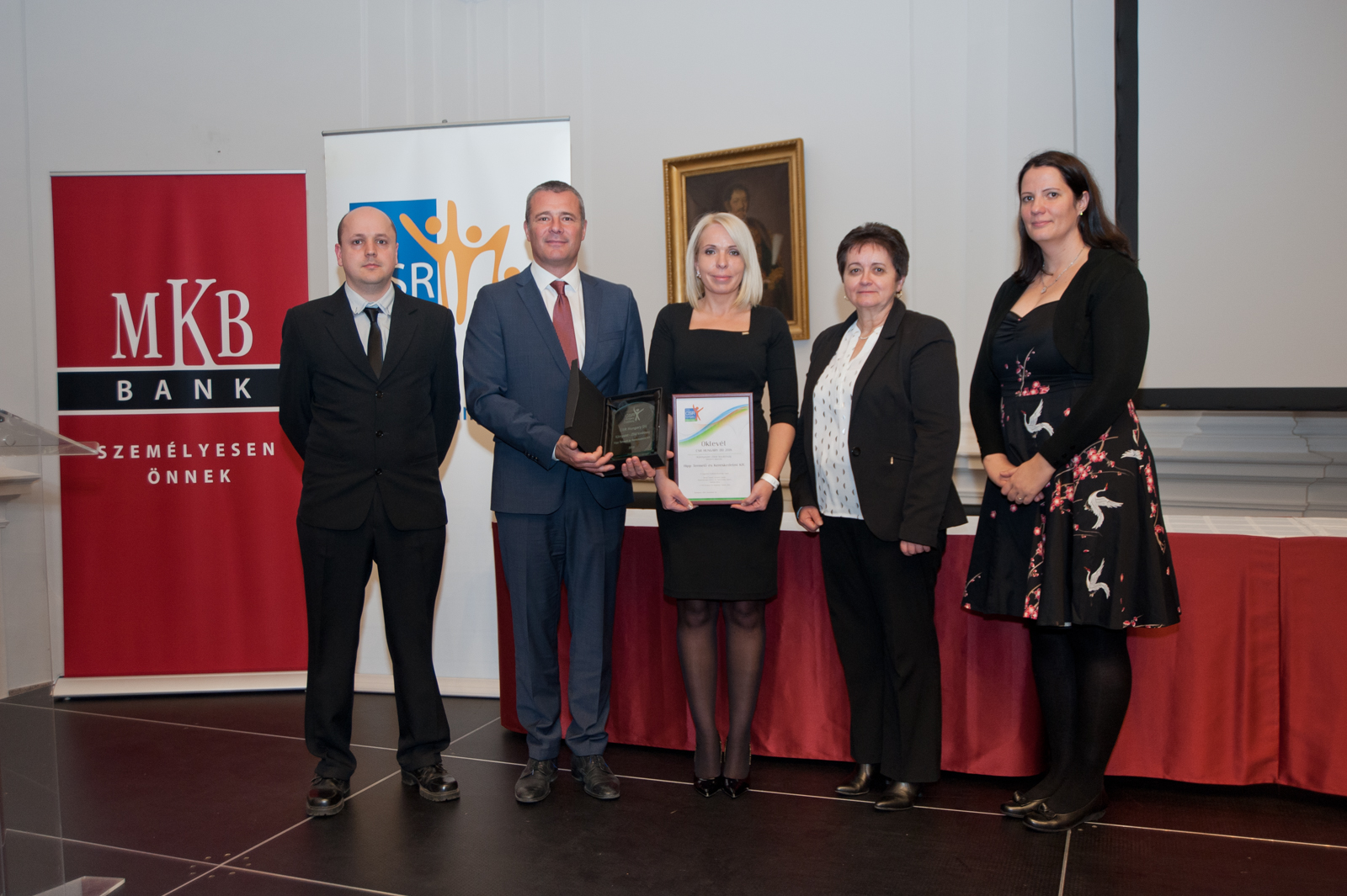 Öt személy áll egy színpadon, ketten egy oklevelet és egy plakettet tartanak a kezükben, egy piros transzparens előtt, amelyen az "MKB Bank" logója látható.